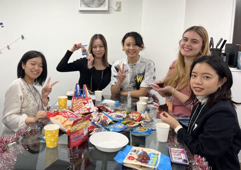 Five women sit together at a glass table covered with snacks, smiling and posing for the camera in an office.