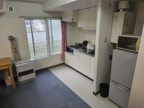 A compact kitchenette with white cabinets and appliances is situated next to a carpeted area with a space heater by a window.