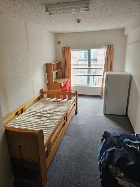 A small dorm room contains a wooden bed, a desk with a red chair, and a white refrigerator next to a window.