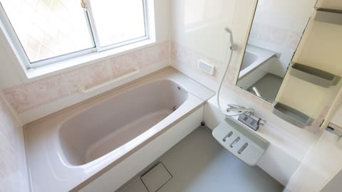 A light-colored bathtub is positioned under a window in a tiled bathroom, with a shower and mirror to the right.