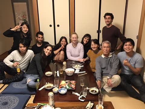 A group of eleven people smiles for a photo while gathered around a low table with food in a room with tatami mats.