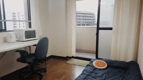A room with a desk, chair, and a bed on the floor, with a sliding glass door offering a view of nearby buildings.