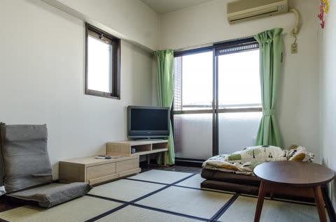 A Japanese-style room with tatami mats, a low TV stand, a floor chair, and a folded futon near a sliding glass door.