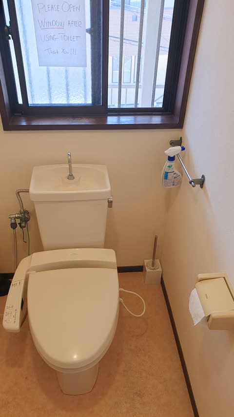 A Japanese toilet with a bidet seat and a sink on its tank sits in a bathroom below a window with a handwritten sign.