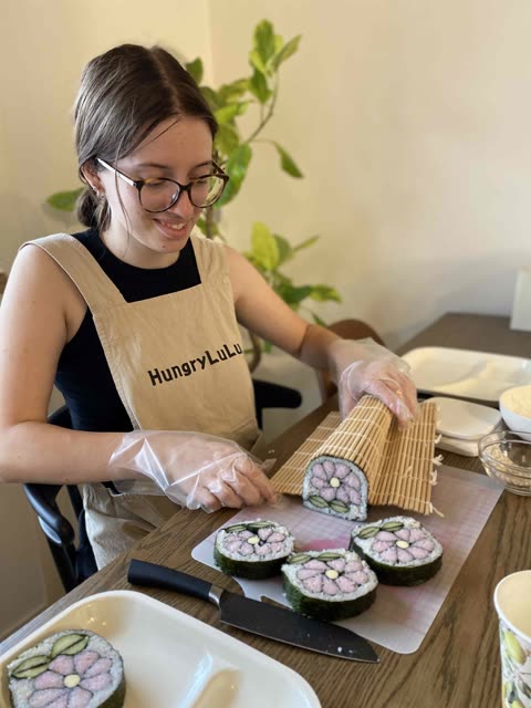 Person wearing apron and gloves makes flower-shaped sushi rolls using a bamboo mat at a table.