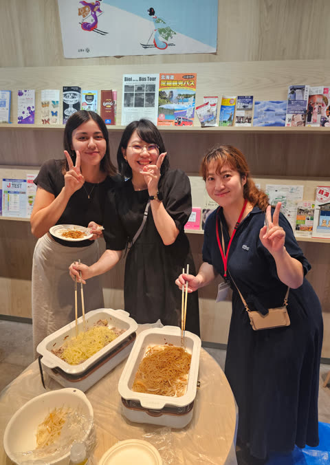 Three women pose with chopsticks near electric skillets of noodles in a room.