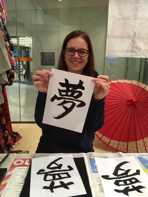 A woman with glasses holds up a piece of paper with a Japanese calligraphy character in a room with a red paper umbrella.
