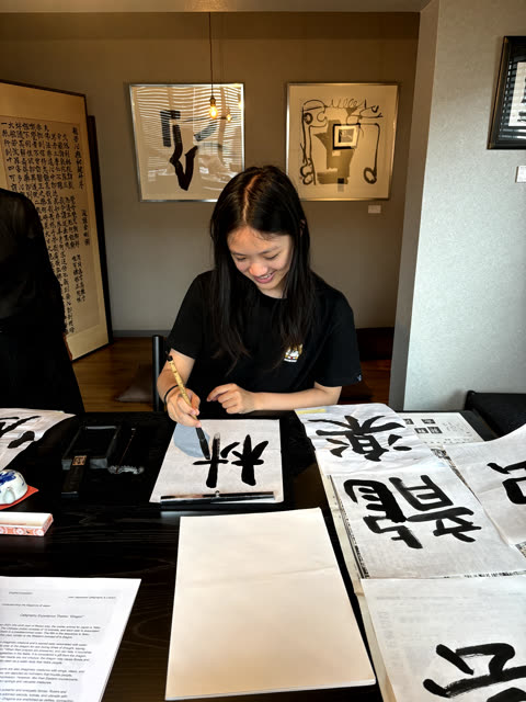 A smiling woman with long dark hair paints a Japanese character with a brush and ink at a table in a studio.