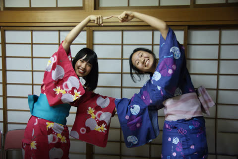 Two women in yukatas form a shape with their arms in front of a room divider.