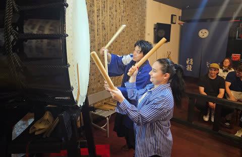 A man in a blue robe and a woman in a striped shirt hold large wooden sticks over a Japanese taiko drum in a room.