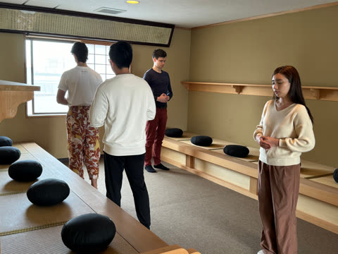 People stand and sit silently in a room with cushions.