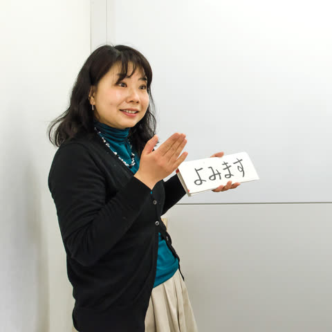 A woman stands in front of a whiteboard holding up a small card with Japanese characters written on it.