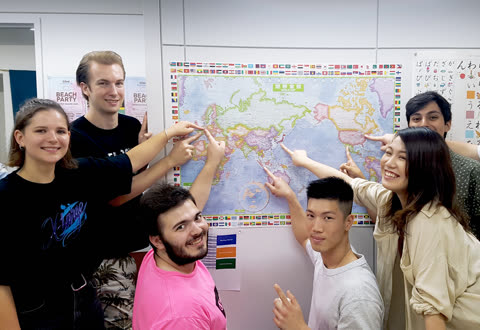 A group of seven people stands in a room, smiling and pointing at different locations on a world map.