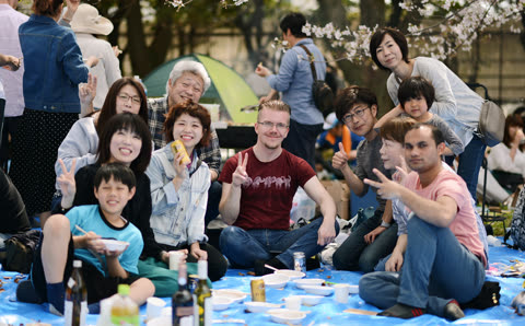A large group of people sits together on a blue tarp in a park, smiling and posing for the camera.