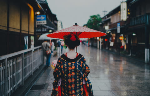 A person from behind in a floral kimono holds a red umbrella while walking down a wet street lined with traditional buildings.
