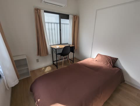 A bedroom with a wood floor contains a bed with brown bedding and a desk with a chair under a window.