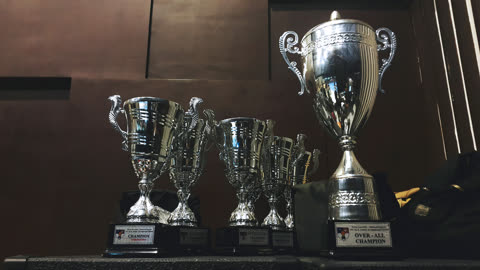 Silver trophies and cups sit on a dark surface against a brown wall.