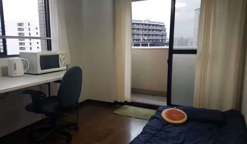 Small room with a bed, desk, microwave, and electric kettle, next to a window showing buildings.