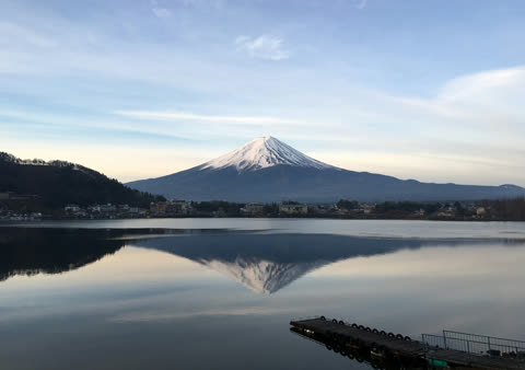 Snow-capped Mount Fuji reflected in a calm lake, with a small dock in the foreground.