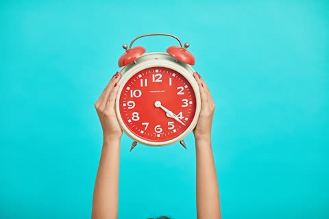 Person holds red alarm clock overhead against a blue background.