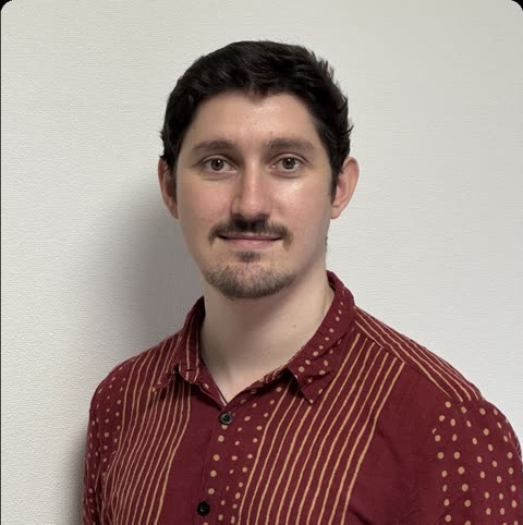 A man with dark hair and a mustache in a red patterned shirt looks at the camera in front of a white wall.