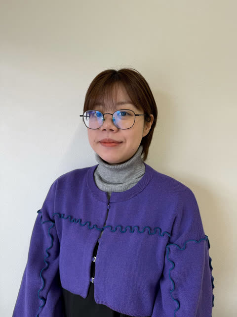 A person with short brown hair and glasses wearing a purple cardigan and a grey turtleneck stands against a plain wall.