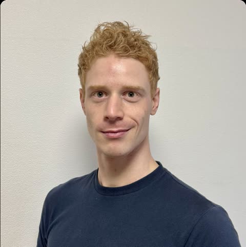 A man with curly red hair and a dark blue t-shirt smiles slightly while looking at the camera against a white wall.