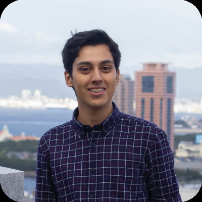 A young man with braces in a plaid shirt smiles on a rooftop overlooking a city skyline and body of water.