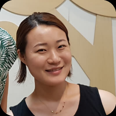 An East Asian woman with shoulder-length brown hair and a black top smiles while standing indoors.