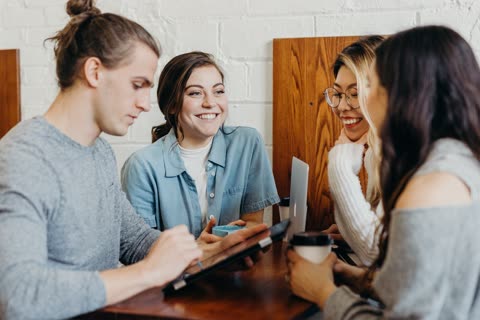 Four people sit at a wooden table in a cafe, smiling while looking at a laptop and a tablet.