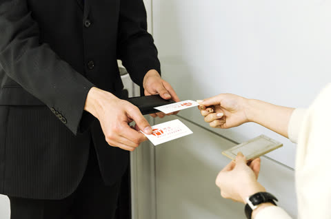 A person in a black suit hands a business card to another person in front of a plain white wall.