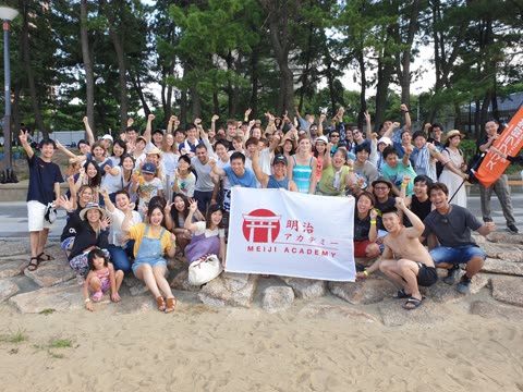 Large group posing with Meiji Academy banner on a sandy beach.